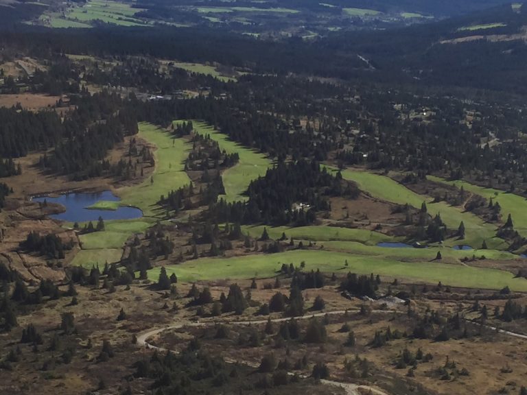 Utsikt over golfbanens fairwayer og vann, omgitt av skog og fjellterreng.
